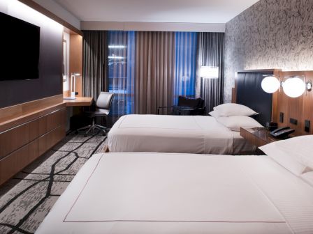 A modern hotel room with two neatly made beds, a wall-mounted TV, desk with chair, and lamps on the headboards.
