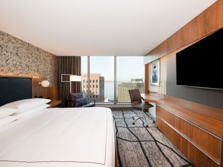 A modern hotel room with a large bed, wall-mounted TV, desk with chair, and a window offering a city view.