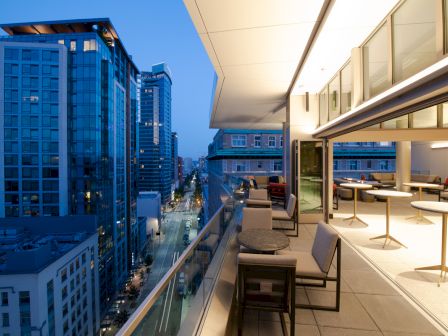This image shows a cityscape from a modern rooftop patio with outdoor seating and tables, overlooking buildings and a street at dusk with building lights on.