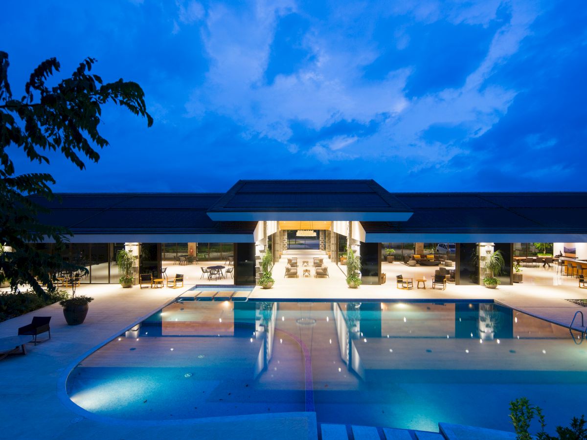 An illuminated modern building with large windows, an outdoor swimming pool, and lounge chairs at dusk, under a blue sky with scattered clouds.