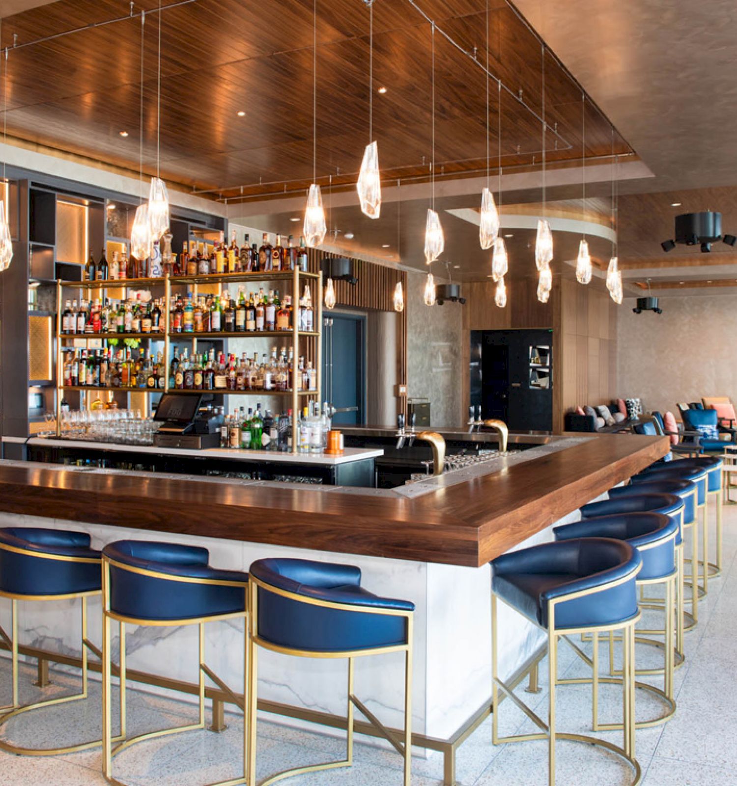 The image shows a modern bar with blue stools, a fully stocked liquor display, pendant lights, and a spacious seating area in the background.