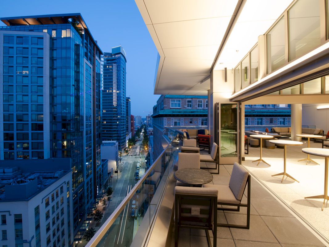A modern cityscape seen from a balcony with outdoor seating, high-rise buildings on both sides, and a well-lit interior area.