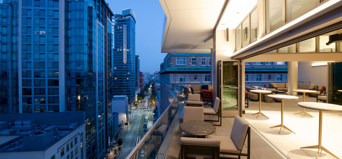 A modern cityscape seen from a balcony with outdoor seating, high-rise buildings on both sides, and a well-lit interior area.