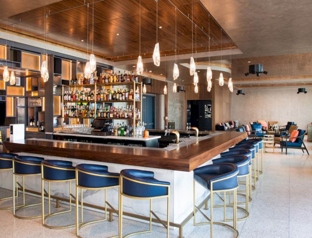 The image shows a modern bar with a wooden counter, blue cushioned bar stools, and shelves of liquor bottles. Elegant pendant lights hang above.