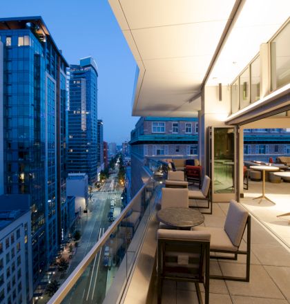 The image shows a modern balcony with seating, overlooking a cityscape filled with tall buildings and streets at dusk.