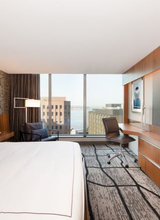 A modern hotel room featuring a large bed, a flat-screen TV, a wooden desk, and a large window with a city view in the background.