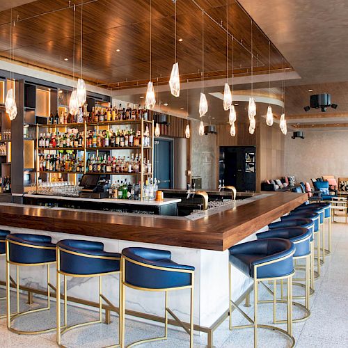 A modern, stylish bar with blue bar stools, hanging light fixtures, a fully stocked liquor shelf, and spacious seating areas in the background.