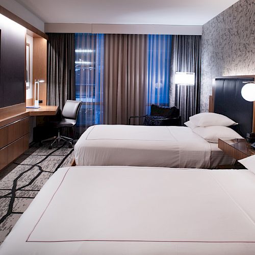 A modern hotel room with two beds, a wall-mounted TV, a work desk with a chair, and floor-to-ceiling windows with curtains.