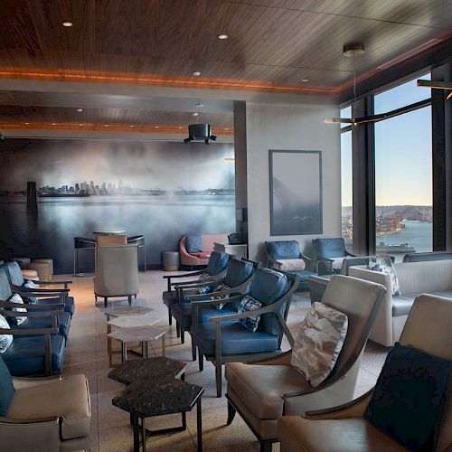 A modern lounge area with plush seating, large windows offering a scenic view of a waterfront and Ferris wheel, and a mural on the back wall.