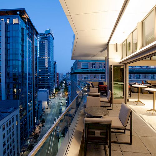An outdoor balcony with seating and tables overlooks a cityscape with tall modern buildings at dusk.