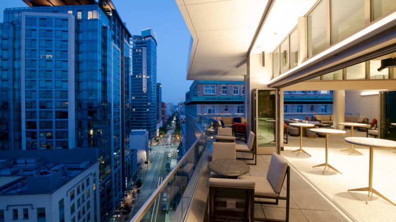 A modern balcony with chairs and tables overlooks a city street lined with tall buildings during evening.