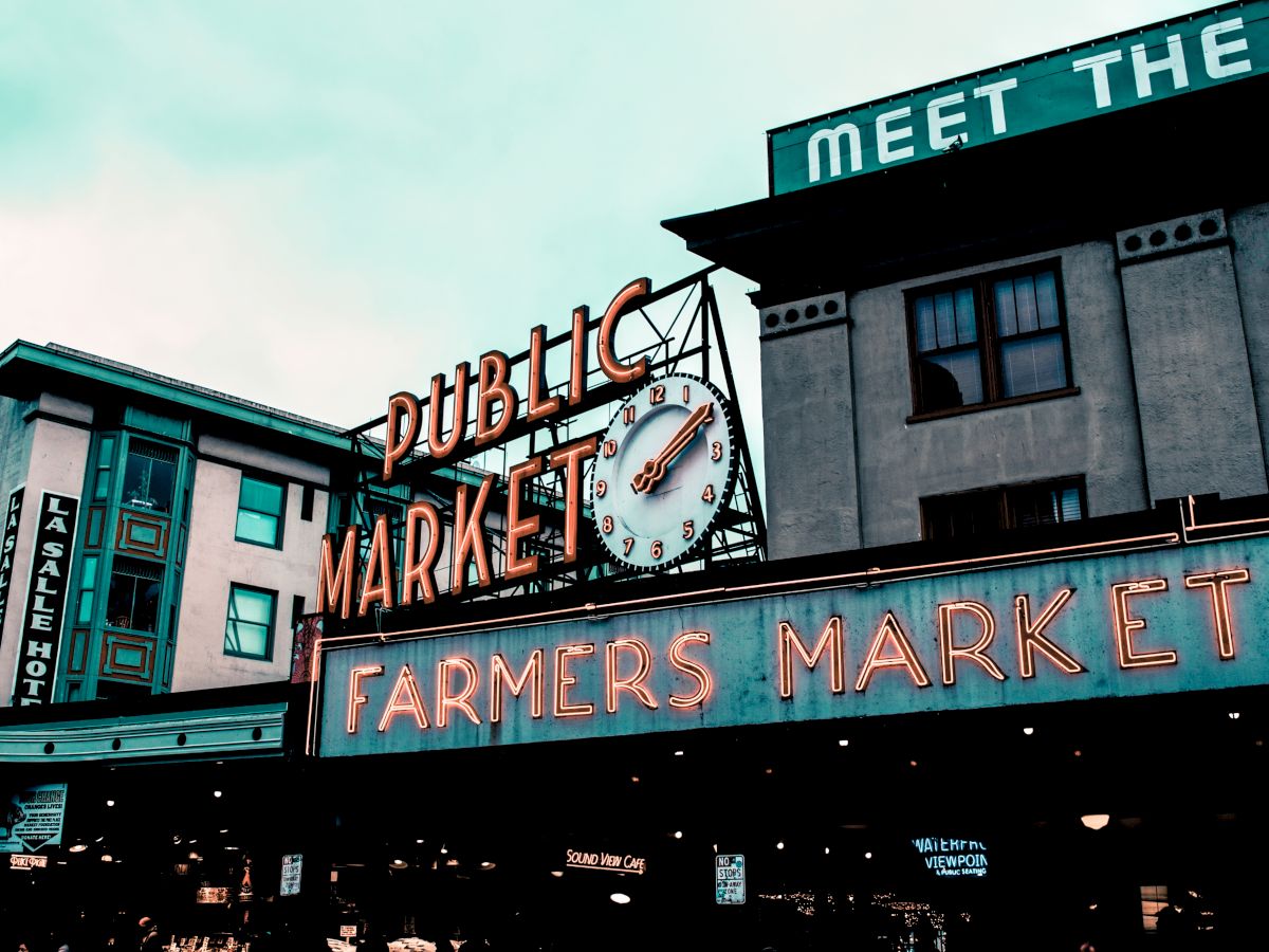 The image depicts an iconic public market sign with "PUBLIC MARKET" and "FARMERS MARKET" prominently displayed in neon lights, with buildings in the background.