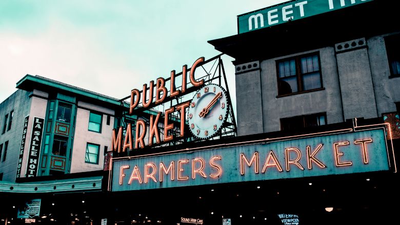 The image depicts an iconic public market sign with "PUBLIC MARKET" and "FARMERS MARKET" prominently displayed in neon lights, with buildings in the background.