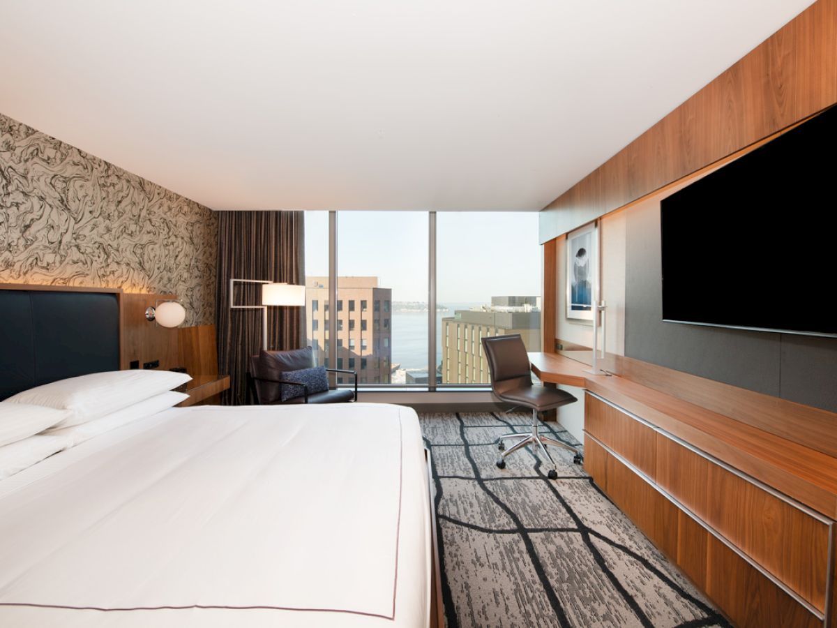 The image shows a modern hotel room with a large bed, wall-mounted TV, desk, chair, and a view of buildings and water through large windows.