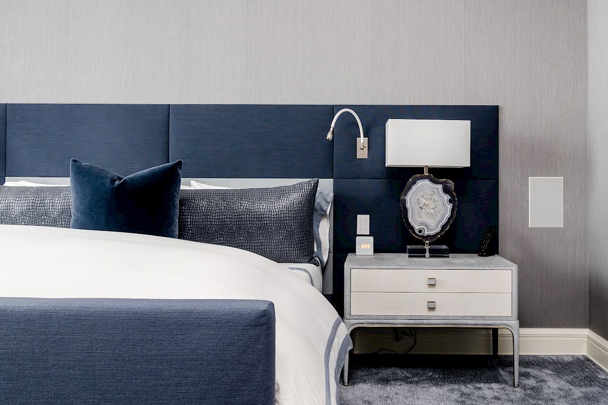A modern bedroom with a blue-upholstered bed, dark blue pillows, a white side table, a decorative lamp, and a textured gray carpet.