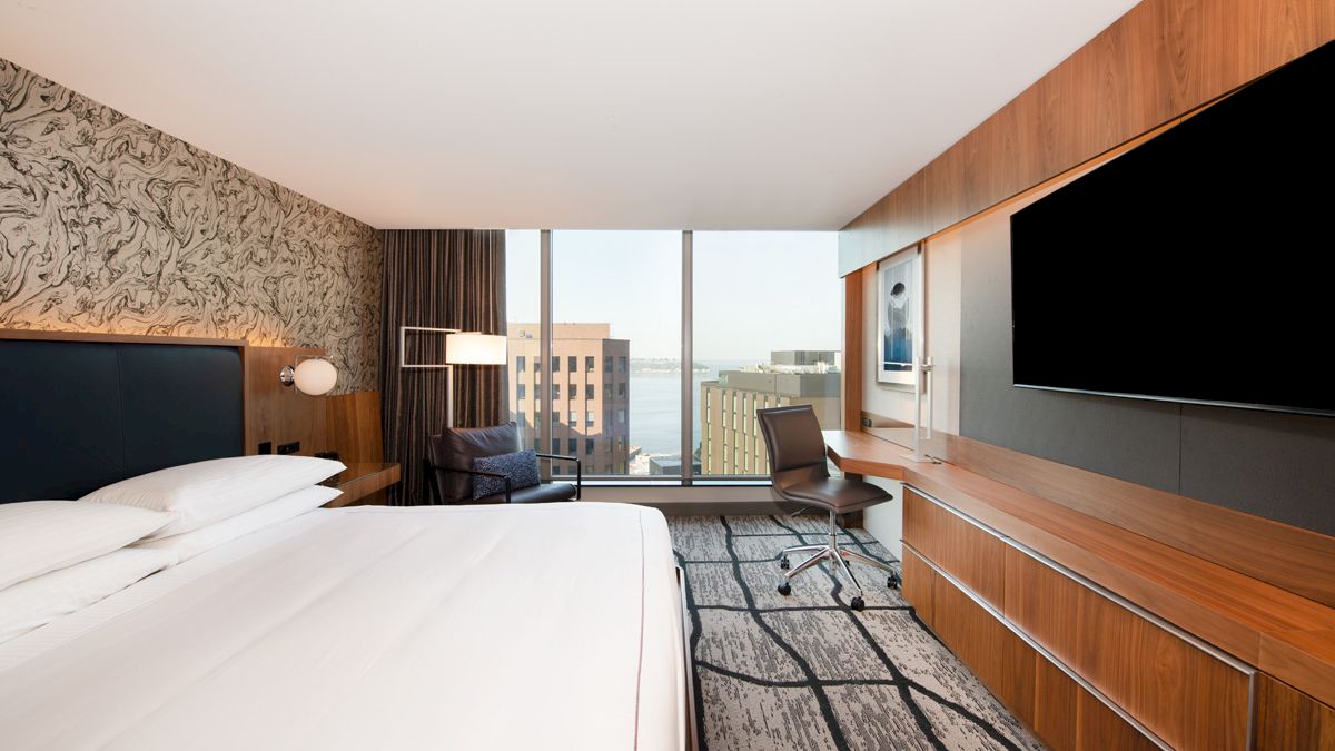 A modern hotel room with a large bed, wall-mounted TV, desk, chair, and large windows offering a city view.