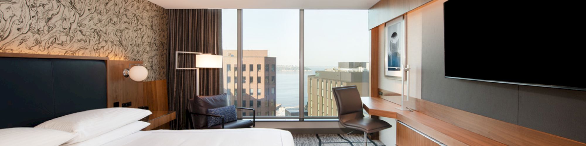 A modern hotel room with a large bed, wall-mounted TV, desk, chair, and large windows offering a city view.